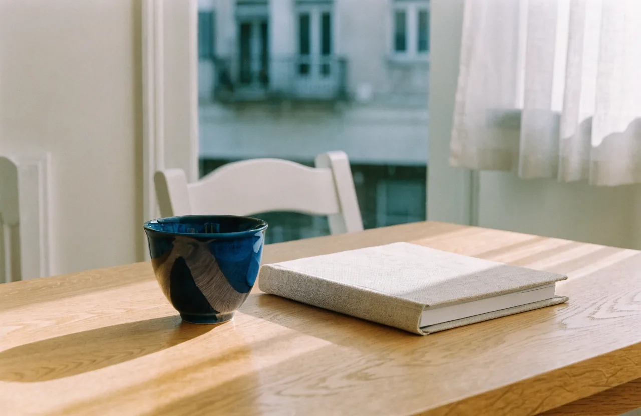 A deep blue glazed ceramic cup beside a closed linen-bound notebook on an oak desk by a window.